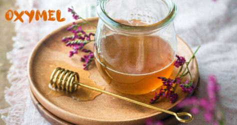 Oxymel - Das Bild zeigt ein Glas mit goldgelber Flüssigkeit und daneben einen Honigportionierer. Oximel ist ein Mix aus Honig, Apfelessiug und Heilkräutern